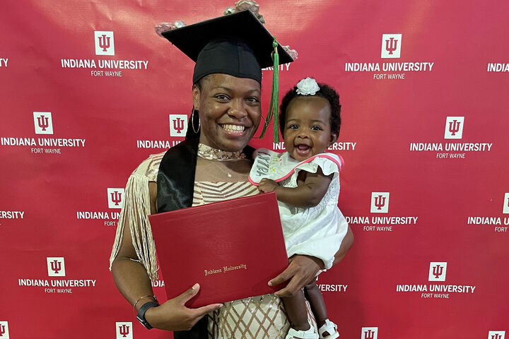 A proud graduate in commencement attire holding her diploma while carrying a happy child.